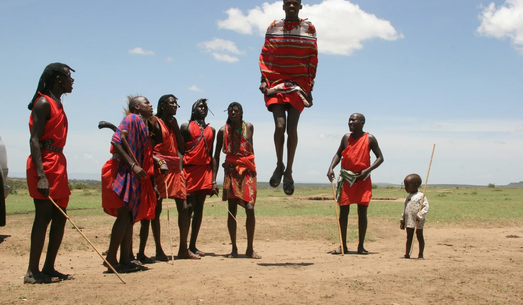 Maasai man in red cloth performing a vertical jump while other men sing in Kenya